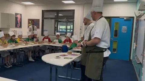 Two professional chefs standing in front of a classroom of children sat down with fresh food and bowls in front of them.