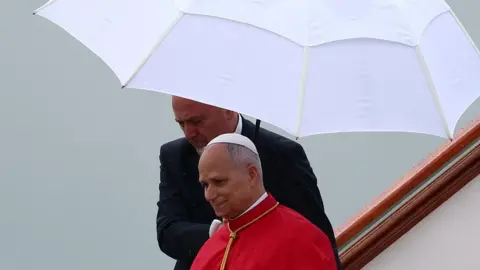 Pope Leo wearing papal cap and robes walks off a plane as a man in a dark suit behind him holds a white parasol over him.