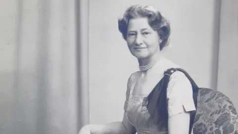 A black and white photo of a lady sat in an arm chair with a long dress on