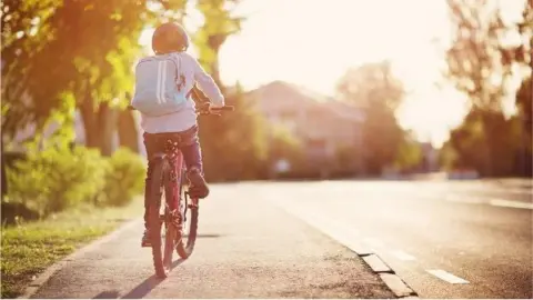 A child wearing a white T-shirt and blue and white rucksack cycles along a pavement. The road is to their right and there is grass with some trees to the left.
