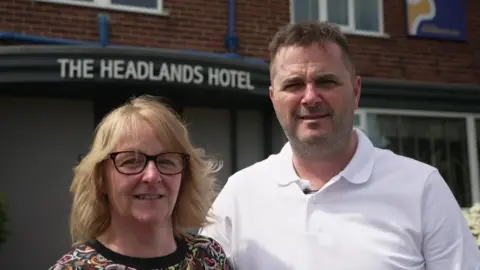 BBC/Luke Manterfield Karen (left) and Steve Ritchie (right) standing outside their hotel in Scarborough. Cathy has blonde hair and glasses and Steve is wearing a white polo-shirt.