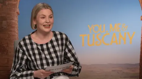 Mared Parry/ Betches Mared sat in front of the You, Me & Tuscany background. She holds a white Betches card with her questions on. She wears a black and white chequed blouse and has short blonde hair.