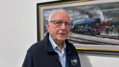 A man with glasses, blue shirt and a blue fleece stood in front of a painting of a train.