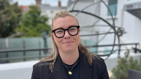 Tourism Advisory Board co-chair Charlie Walker. She is wearing a black top and black blazer. She has long blonde/brown hair which is tied back. She is also wearing thick framed black glasses. She looking at the camera in front of a blurred background of what looks like a hotel outside space.