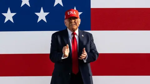 Trump clapping with a huge US flag behind him. He's wearing a suit and a bright red cap that says USA
