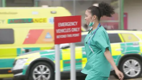 A female medical worker wearing scrubs walks in front of two ambulances. A sign between her and the vehicles reads "emergency vehicles only beyond this point". She has a face mask under her chin and her hair is tied into a ponytail. 
