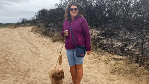 BBC News A woman wearing sunglasses, a purple jacket and denim shorts holds a dog on a lead on the sand. They are standing in front of blackened, burnt trees 
