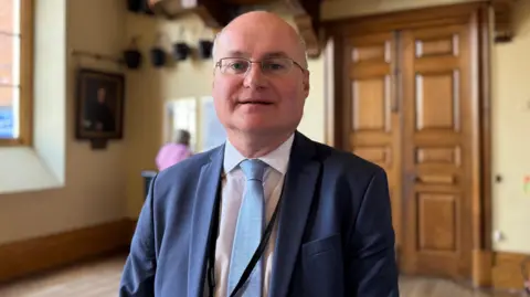 A bespectacled man, bald on top of his head but with grey hair at the sides, is wearing a white shirt with a pale blue tie under a navy blue suit jacket.
