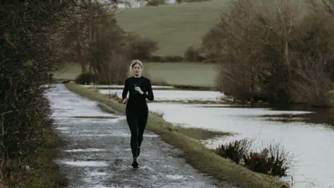 Jack Schofield A young woman runs down the side of a canal. She wears an all black outfit and has her blonde hair tied back. 