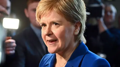 Nicola Sturgeon being interviewed by journalists. She is in close up and is wearing a royal blue blazer