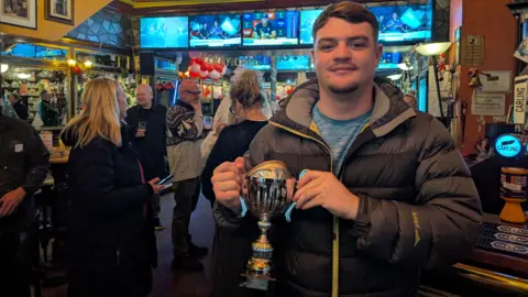 Tom O'Neil stands in a pub with a silver trophy which has a pie inside it. He wears a grey coat and a blue t-shirt and there are some people in the pub behind him.
