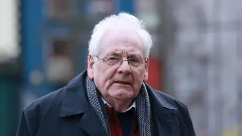 PA Media Michael Gallagher has grey hair and is wearing metal-framed glasses. He is wearing a navy overcoat, grey scarf, red gilet and a shirt and jumper.