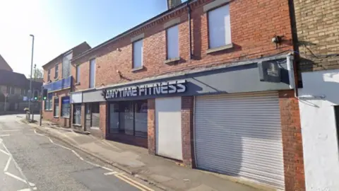 Google A long brick terrace with "Anytime Fitness" written above a doorway. A shutter is down on the shop front next to it.