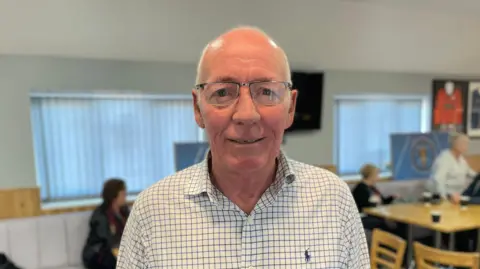 Mark Ansell / BBC A man with bald head and grey hair at the sides, wearing metal-rimmed glasses and a white shirt with thin blue and black checked pattern.