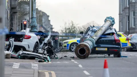 There is a wreck of a white car. Its roof has been removed by the fire service. Near to the car is the other car, partly crushed by an old ventilation shaft. There is debris on the road around the cars. Parked across the road is a police car.