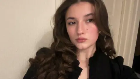 A brunette girl with curly hair, looking towards the camera with a straight expression. She wears a black open blouse and stands against a plain white back drop.