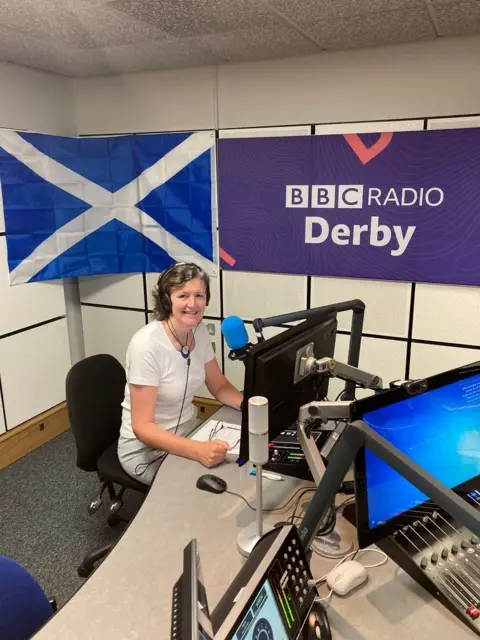A woman sat at a microphone with a Scotland flag on the wall. BBC Radio Derby branding is also visible. 