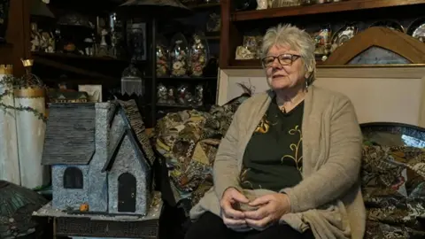 Jayne sat in her house surrounded by the items she collects, including ornamental cats and framed pictures. She has glasses, short grey hair and is wearing a light brown cardigan over a green patterned top.