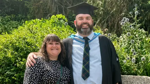 David Spelman and a woman. He has a grey and dark beard and is wearing a graduation gown. The woman has long brown hair and is wearing a black an white patterned dress. 