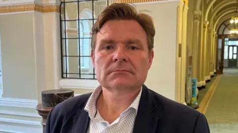 BBC A man wearing a shirt and dark jacket is standing in a corridor. A green carpet and a door in the background on the right are visible. He has brown hair with a side parting.

