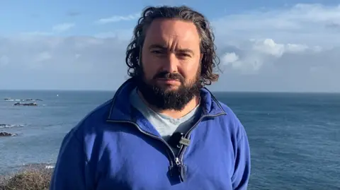 Lee van Katwyk is looking serious as he stands with the sea behind him on a bright blue sky day. He has shoulder-length dark curly hair and a dark beard and moustache. He is wearing a blue half-zipped sweatshirt over a pale blue top and has a microphone clipped to his collar. 