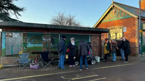 A group of people in thick coats and hats stand outside the bus shelter which has been fenced off