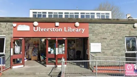 A general view of the entrance to Ulverston Library. The one-storey grey building has a red sign with white lettering on top of the door, identifying it as the library. 