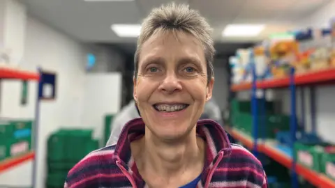 Lucy Martin is smiling at the camera, has blue eyes and short grey hair. She is wearing a pink and purple fleece and in the background are shelves and crates for food.