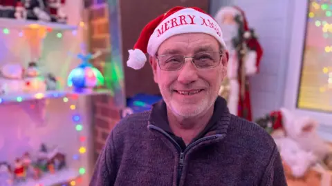 BBC/Jodie Halford A man wearing glasses and a Santa hat smiles at the camera. Multicoloured fairy lights are visible in the background 