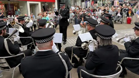 A brass band performance in front of an audience taking place on a pedestrianised city street. In the foreground musicians are seated in a semi-circle facing a conductor. They are dressed in dark formal uniforms with matching caps and are sitting on chairs holding brass instruments.