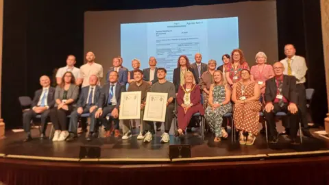 Redditch Borough Council A large group of people, including Jordan and Cian Adams are sat across two rows of seats on a stage, with a projector behind them. 