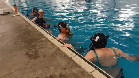 Fiona Callow/BBC A mixed gender group of people are in the water by the side of a swimming pool. Everybody wears black snorkels, swimming caps and masks.