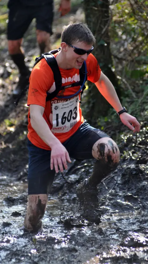 Photofit.com A man attempts to run through a bog during a race in Devon called The Grizzly. He is wearing dark shorts, a yellow t-shirt and a black backpack. He has a number on the front of his shirt with 1603 on it. He is knee-deep in mud.