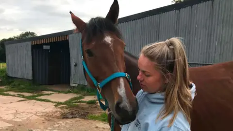 Hayley Vaughan A blonde white woman with her hair in a ponytail wearing with a grey hooded sweater holds a brown horse with a white patch on its nose close to her. Behind them is a yard with a grey corrugated building with a black door 