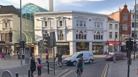 A McDonald's shop front at a busy junction with a small van and two cars, two cyclists and pedestrians at a pelican crossing.