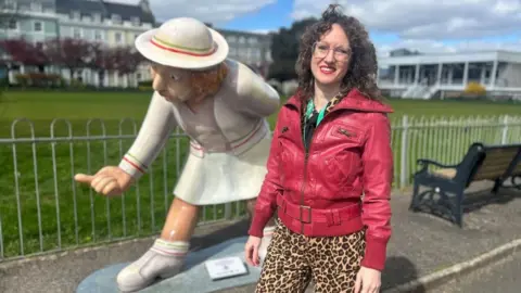 Terah has long brown curly hair, brown leopardskin trousers and a red leather jacket. She is standing in front of a statue of a woman bowler in front of a bowling green