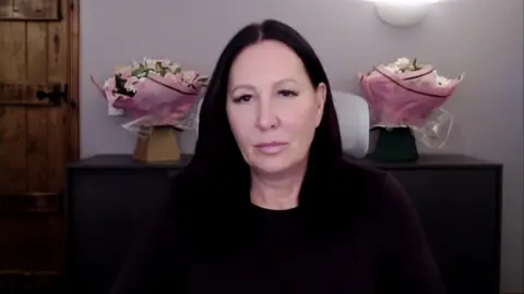 A dark-haired woman in dark clothes framed centre with two pink flower bouquets either side of her head