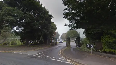 The entrance to Shiregreen Cemetery in Sheffield. It has a large stone gateway, with trees and iron railings around the perimeter.