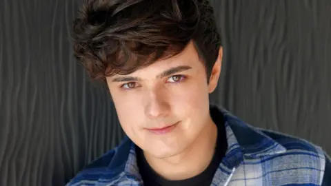 Crancerol Jack Agillor, un joven con pequeño cabello negro, está parado frente a un fondo gris con risas en la cámara para este tiro en la cabeza de estudio.