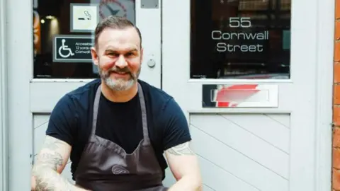 Claire Lishman PR A man with short, dark hair and a beard, is wearing a black T-shirt and a grey apron, sits on the steps outside a building. One of the glass doors is labelled with "55 Cornwall Street".