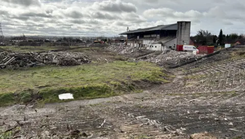 The site of Casement Park in west Belfast. There is rubble on the main pitch. There are terraces to the right.
