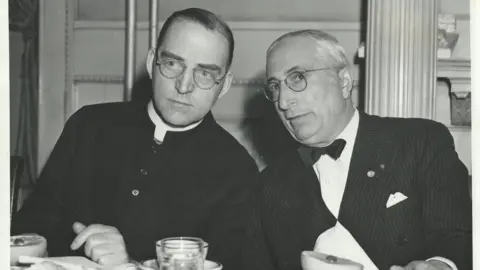 Boys Town Archives Two men sit side by side at a dinner table. The image is black and white and appears to be taken some time ago. The man on the left is wearing clerical garb and the man on the right is formally dressed, including a dinner jacket and bow tie. Both men are wearing glasses.