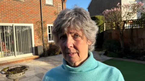 Martin Heath/BBC Penny Holton with medium-length white hair, wearing a blue pullover and standing on the lawn outside a brick-built house with vertical blinds across the patio doors. There is a stone bird bath to her right.