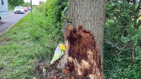 The damaged tree, with grass around it and a boquet of flowers laid beside it