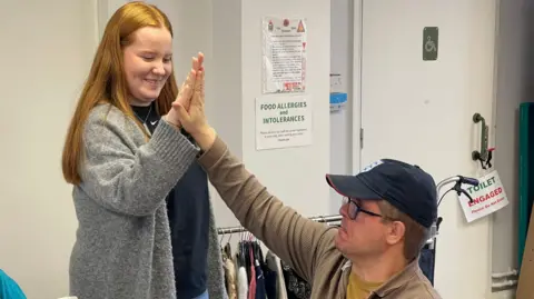 A woman with long red hair wearing a long, grey, thick woollen cardigan gives a high five to a man who is sat down in a chair. He is wearing an England baseball cap, a brown jacket and glasses.