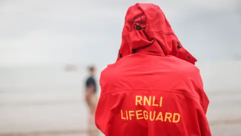 A picture of a lifeguard standing on a beach wearing a large red coat. It is a red coat with yellow writing on it reading: RNLI LIFEGUARD.
