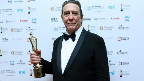 Charles McQuillan/Getty Images Ciarán Hinds, a man with greying dark hair, holds his Ifta award in front of a white wall covered in sponsors' logos. His hair is slicked back and he is wearing a black tuxedo, a white shirt and a black bowtie.