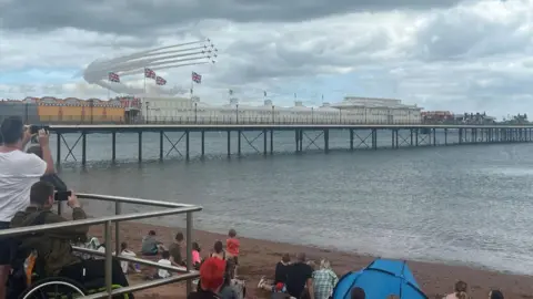 Crowds of people watch on from the beach as the RAF Red Arrows fly in formation over Paignton Pier on a cloudy day. Smoke is coming out of the rear of the aircraft.
