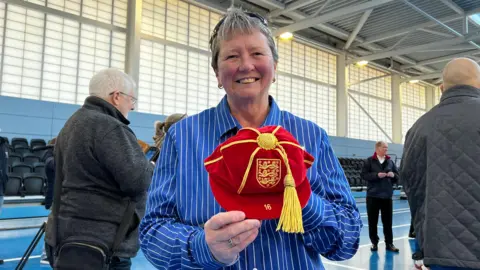A smiling Pat Mitchell-Firth wears a blue and white striped shirt and is holding a crimson velvet official Football Association cap with gold embroidery and brocade. It bears the number 16 and has the England three lions badge on
