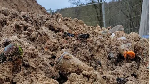 National Trust Picture of a mound of sand with lots of rubbish mixed in. In the photo there is broken glass, rusty tines and bottles